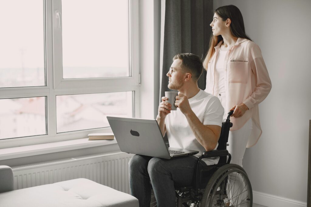 Man in wheelchair with laptop and coffee, gazing out window with a woman beside him.